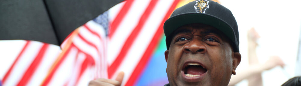 Ras Baraka addressing the crowd at the Newark No Kings rally in the rain, US and Pride flag flying behind him
