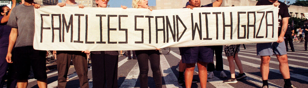 35mm color photo of protestors holding a banner reading "Families Stand With Gaza." They're blocking the street in front of the Brooklyn Library. New York State Assembly member Marcela Mitaynes is one of the protestors, holding up her fist. Organizer Eon Huntley holds a corner of the banner on the far left.
