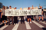 35mm color photo of protestors holding a banner reading "Families Stand With Gaza." They're blocking the street in front of the Brooklyn Library. New York State Assembly member Marcela Mitaynes is one of the protestors, holding up her fist. Organizer Eon Huntley holds a corner of the banner on the far left.