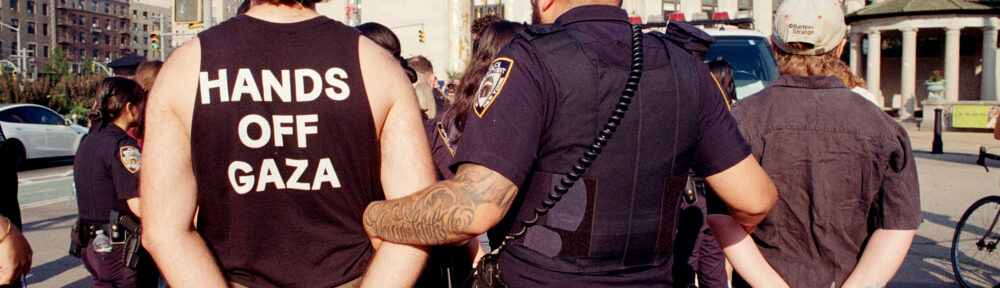 A tattooed cop walks away two ziptied protestors at the "Families Stand Up for Gaza" rally. We're looking at them from behind. One of the protestors is wearing a "HANDS OFF GAZA" t-shirt. 35mm color photo. Brooklyn Public Library in the background.