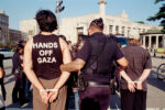 A tattooed cop walks away two ziptied protestors at the "Families Stand Up for Gaza" rally. We're looking at them from behind. One of the protestors is wearing a "HANDS OFF GAZA" t-shirt. 35mm color photo. Brooklyn Public Library in the background.