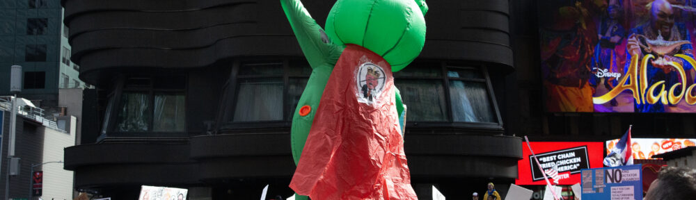 Person in an inflatable frog costume with a cape and crown strikes a John Travolta disco pose while standing on a row of garbage cans above the crowd at the Manhattan No Kings protest.