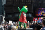 Person in an inflatable frog costume with a cape and crown strikes a John Travolta disco pose while standing on a row of garbage cans above the crowd at the Manhattan No Kings protest.