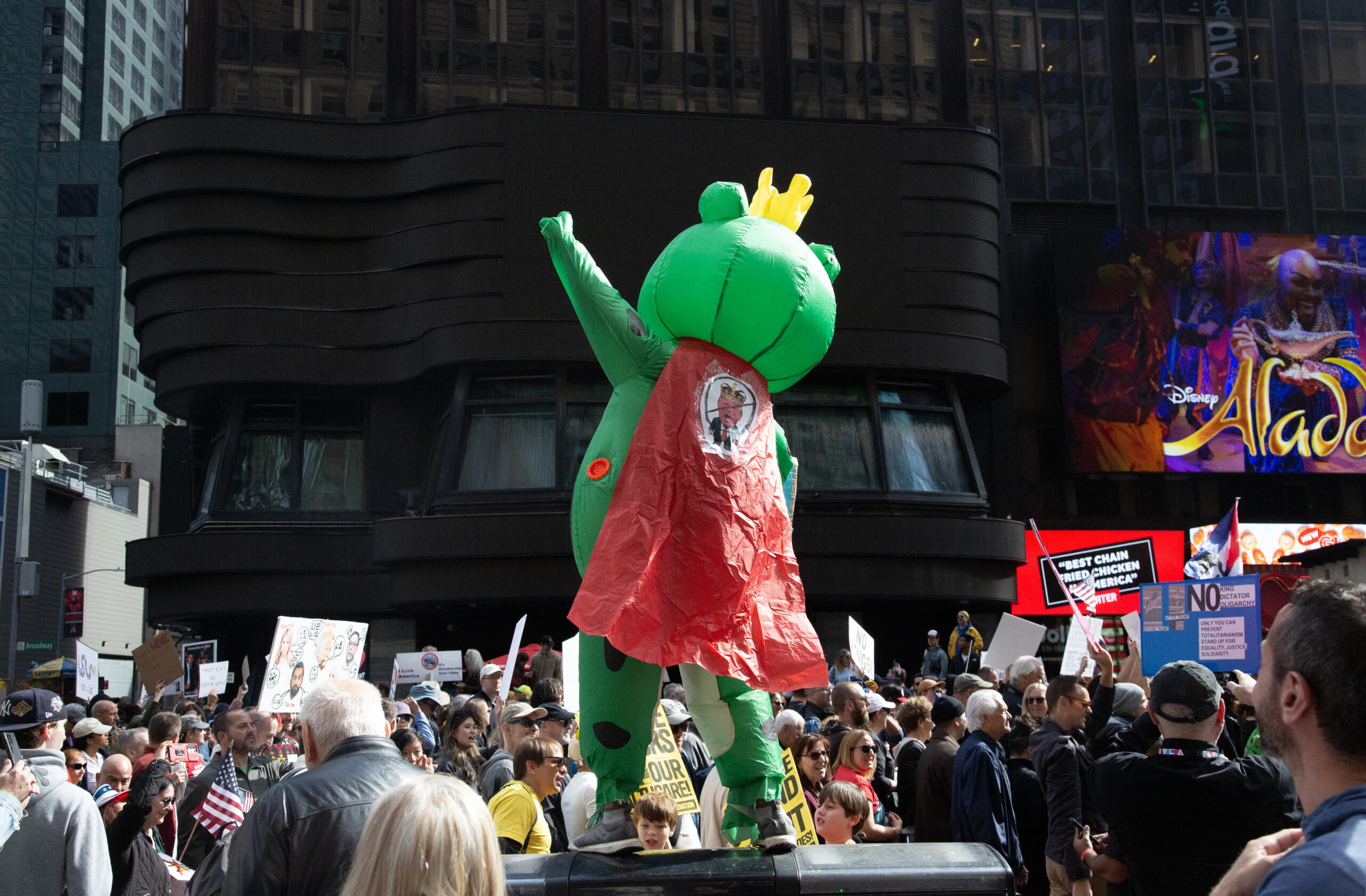Person in an inflatable frog costume with a cape and crown strikes a John Travolta disco pose while standing on a row of garbage cans above the crowd at the Manhattan No Kings protest.