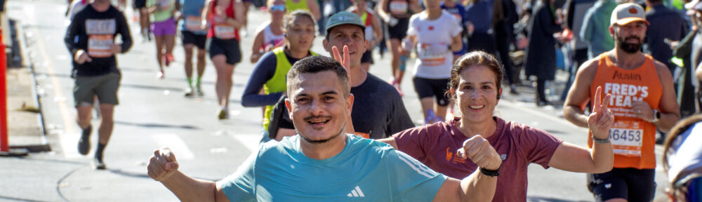 Two runners approach the camera at the New York marathon - both holding up hands, one making fists; the other making "V" signs. Both look really happy. Hundreds of other runners behind them, all backlit. Pretty lovely!