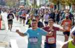 Two runners approach the camera at the New York marathon - both holding up hands, one making fists; the other making "V" signs. Both look really happy. Hundreds of other runners behind them, all backlit. Pretty lovely!