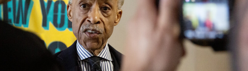 Rev. Al Sharpton getting interviewed after the press conference. We're looking at him over the shoulders of the interviewer on the left and a videographer on the right - screen of the video camera blurry in the foreground.
