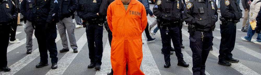 A person in a Trump mask wearing an orange prisoner's uniform stands before a bunch of cops at the end of the No Kings march in Manhattan. Effect is to make it look like they've arrested him. The cops are looking off in other directions, trying to ignore the person.