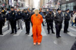 A person in a Trump mask wearing an orange prisoner's uniform stands before a bunch of cops at the end of the No Kings march in Manhattan. Effect is to make it look like they've arrested him. The cops are looking off in other directions, trying to ignore the person.