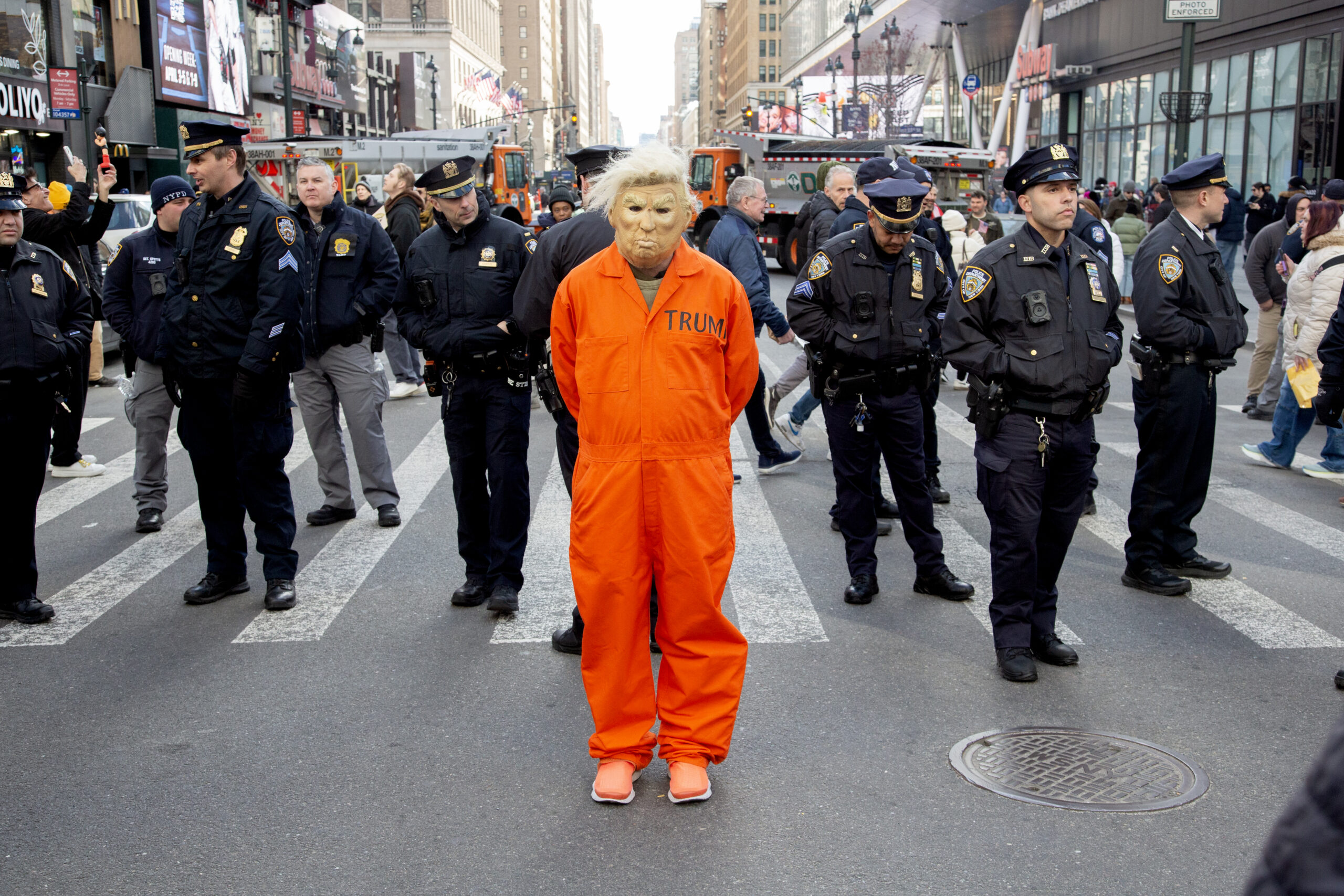 A person in a Trump mask wearing an orange prisoner's uniform stands before a bunch of cops at the end of the No Kings march in Manhattan. Effect is to make it look like they've arrested him. The cops are looking off in other directions, trying to ignore the person.