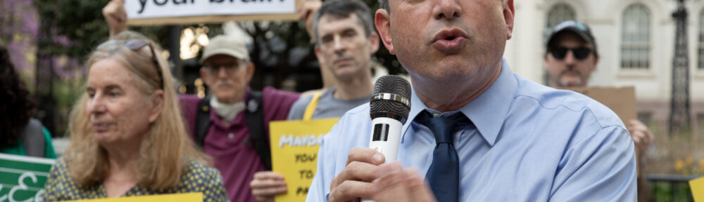 Brad Lander gestures with palm facing upwards at a rally in front of City Hall. Person in the background holds a sign reading "AI rots your brain"