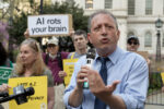 Brad Lander gestures with palm facing upwards at a rally in front of City Hall. Person in the background holds a sign reading "AI rots your brain"