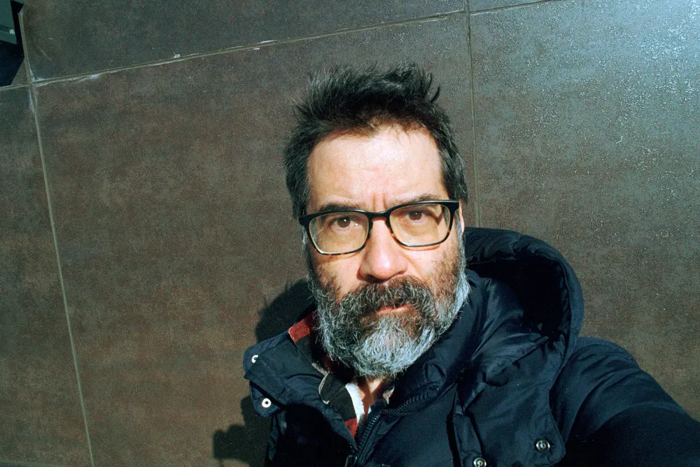Selfie of Greg Pak taken on 35mm film in the winter in 35mm color. He's standing against a stone wall, bundled up in a puffy coat. Grizzled, with glasses.