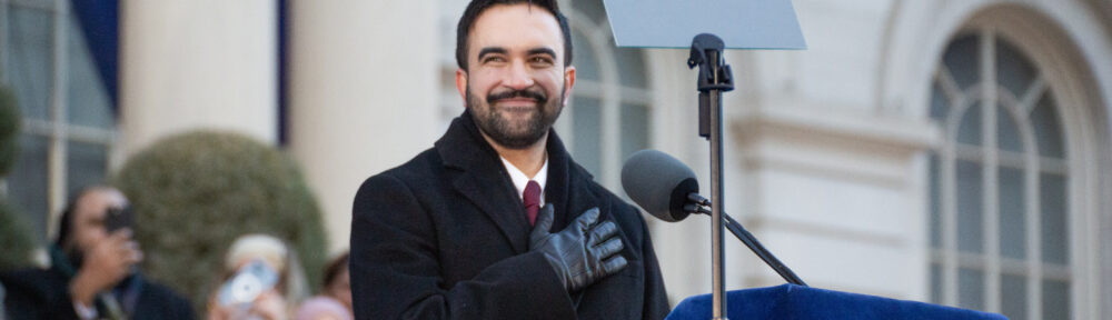 Mamdani smiles, hand to his heart, as he stands at the podium during his inauguration.
