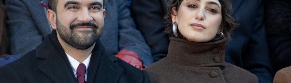 Zohran Mamdani and Rama Duwaji sitting at the inauguration. He's smiling. She's gazing towards the camera with her head tilted and a look of quiet regard? judgement? amusement? on her face.