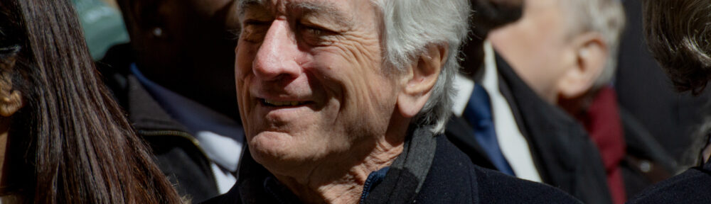 Robert De Niro smiles while looking off to the side while marching in No Kings Manhattan. He's wearing a button that says "I [heart] IMMIGRANT NY"