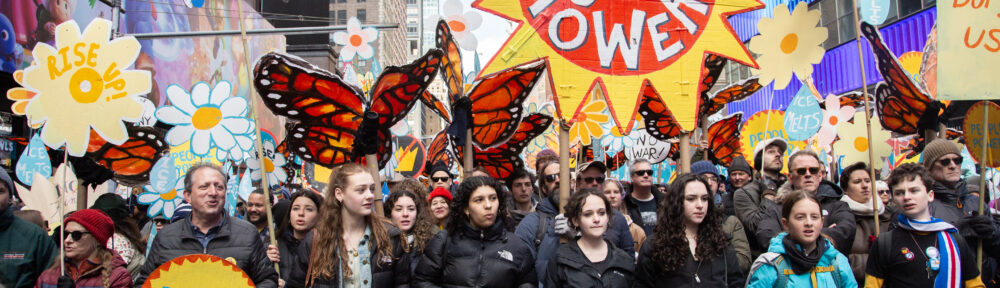 Indivisible Brooklyn members marching with Brad Lander during No Kings Manhattan behind a banner reading "TOGETHER WE RISE." Gorgeous art placards, one with a sun reading "WE ARE THE POWER," others with monarch butterflies and flowers. Lander's carrying a placard reading "PEOPLE POWER"