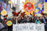 Indivisible Brooklyn members marching with Brad Lander during No Kings Manhattan behind a banner reading "TOGETHER WE RISE." Gorgeous art placards, one with a sun reading "WE ARE THE POWER," others with monarch butterflies and flowers. Lander's carrying a placard reading "PEOPLE POWER"
